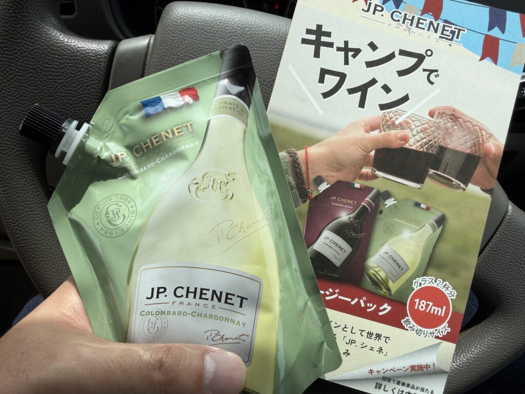 車内で手に持ったパウチタイプの白ワイン「JP CHENET」と、キャンペーン案内のチラシが写っている様子