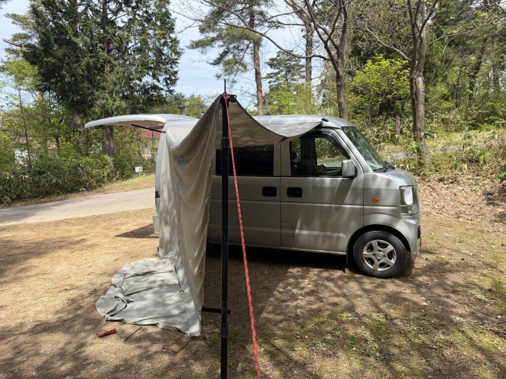 林間のキャンプサイトで軽バンの横にカーサイドタープを設営している様子