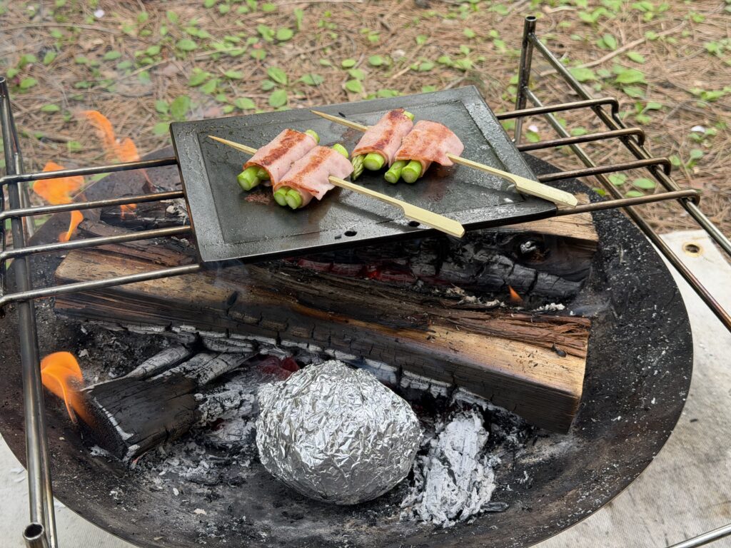 焚き火台の上でアスパラベーコンを焼きながら、下の熾火で新玉ねぎのホイル焼きをしている様子