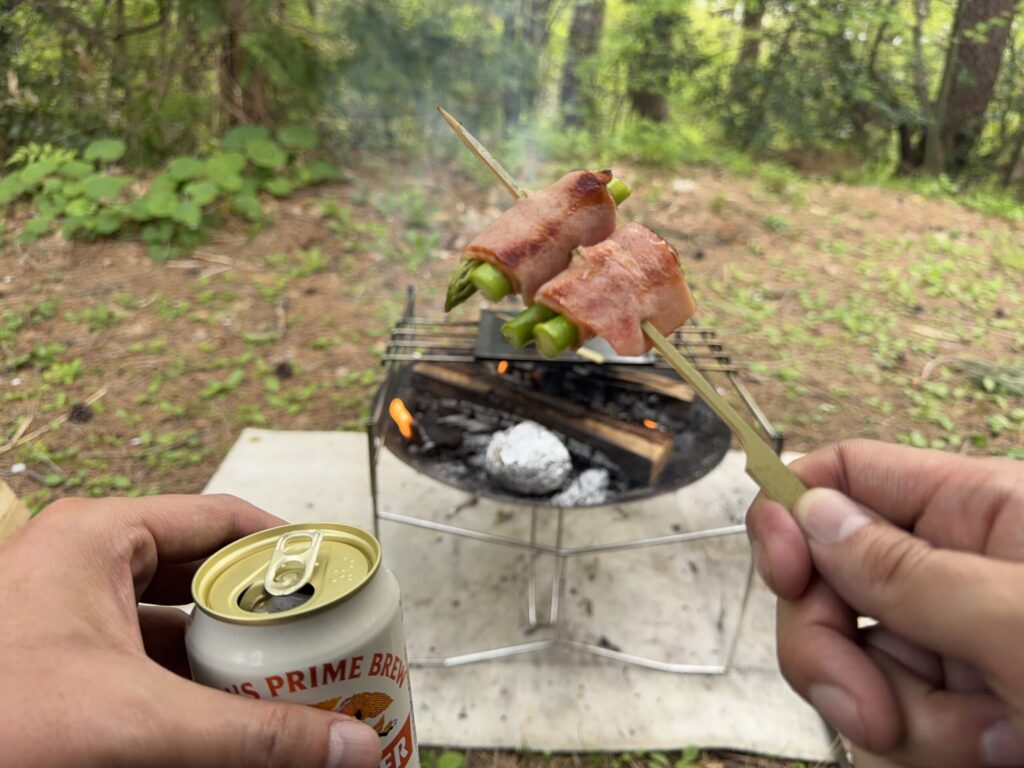 焚き火台を背景に、手に持ったビールと焼き上がったアスパラベーコンを食べている様子
