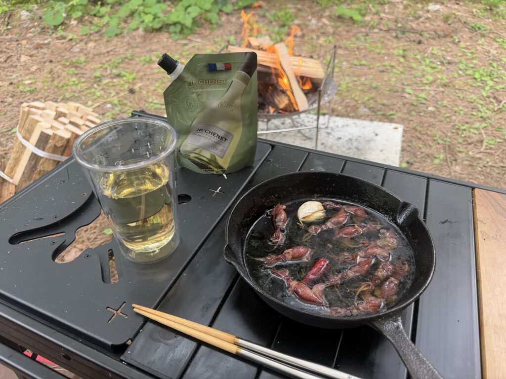 スキレットに入ったホタルイカのアヒージョと白ワインのグラスがテーブルに置かれ、焚き火を背景にしている様子