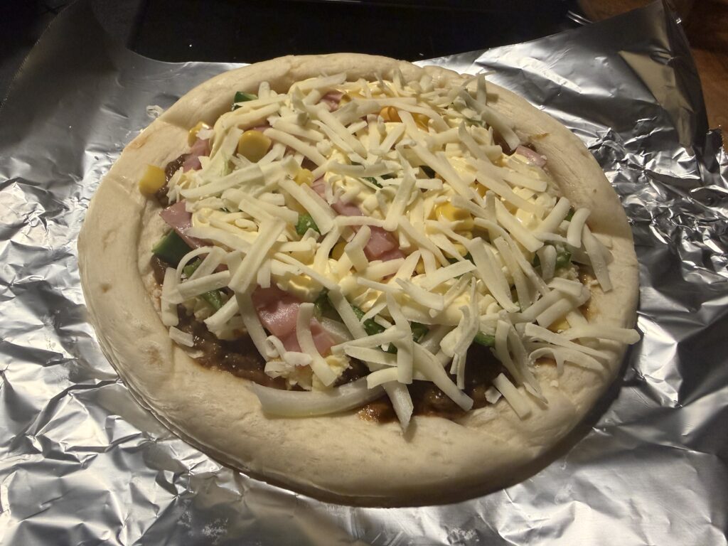 アルミホイルの上に置いたピザ生地に、カレーソース、ポテトサラダ、ベーコン、ピーマン、コーン、チーズを乗せた焼く前の様子。