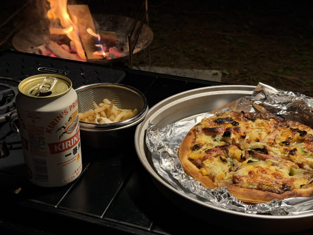 キャプテンスタッグのピザグリルで焼き上げたカレーモントレー風ピザとビールを焚き火のそばに並べた様子