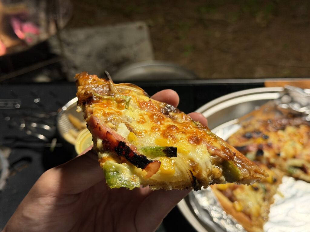 焼き上がったカレーモントレー風ピザを一切れ手に持ち、キャンプ場で食べようとしている様子