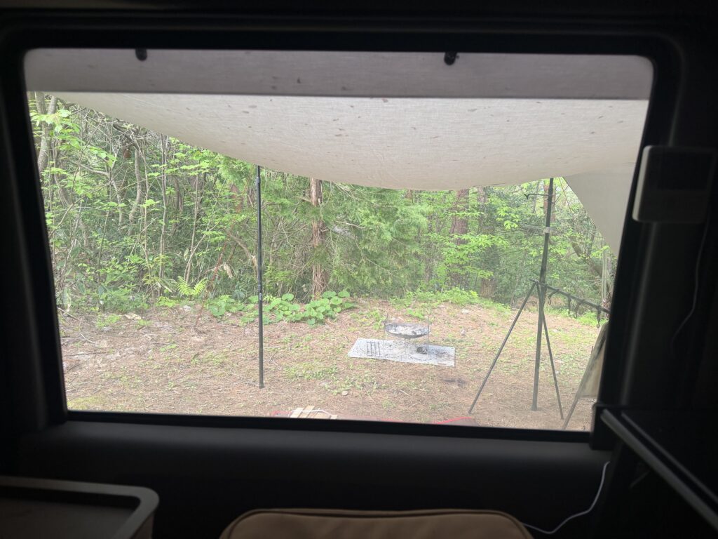 朝起きて車内のサンシェードを外し、窓越しにキャンプサイトの森と焚き火台を眺めている様子