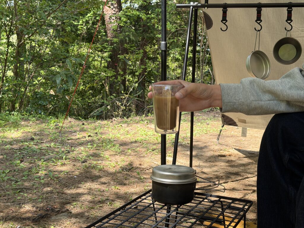 キャンプ場のタープ下で米を浸水させながら、手に持ったアイスコーヒーを飲んでいる様子