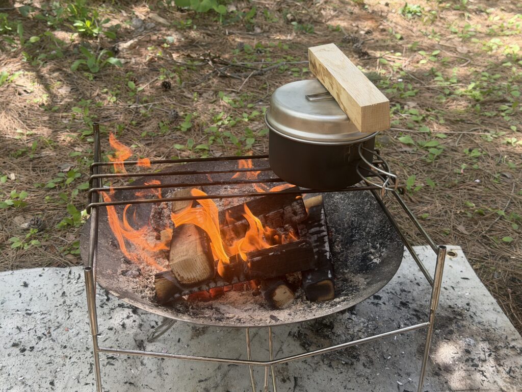 キャンプ場で焚き火を使い、クッカーに木を重しとして乗せて水蒸気炊飯している様子