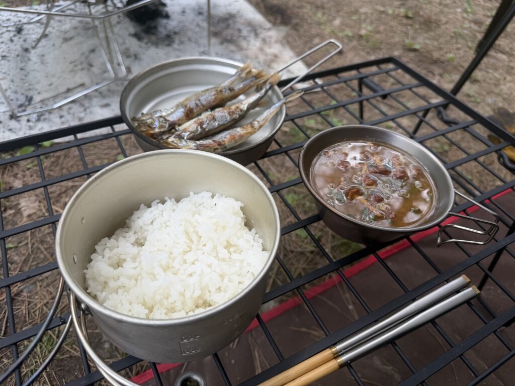 キャンプ場のテーブルに炊きたてのごはん、熾火で焼いたししゃも、インスタント味噌汁を並べた昼ごはんの様子