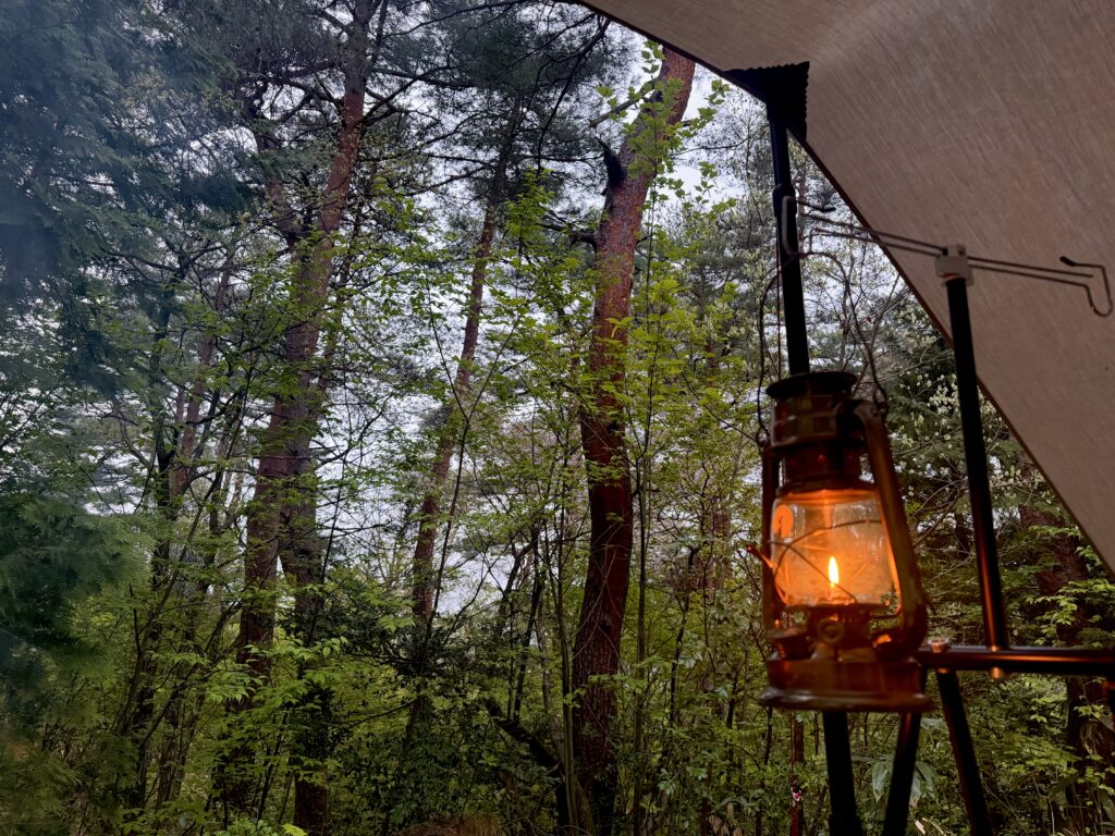 カーサイドタープの下でオイルランタンに火を灯し、周囲の林が夕暮れで少し暗くなってきた様子