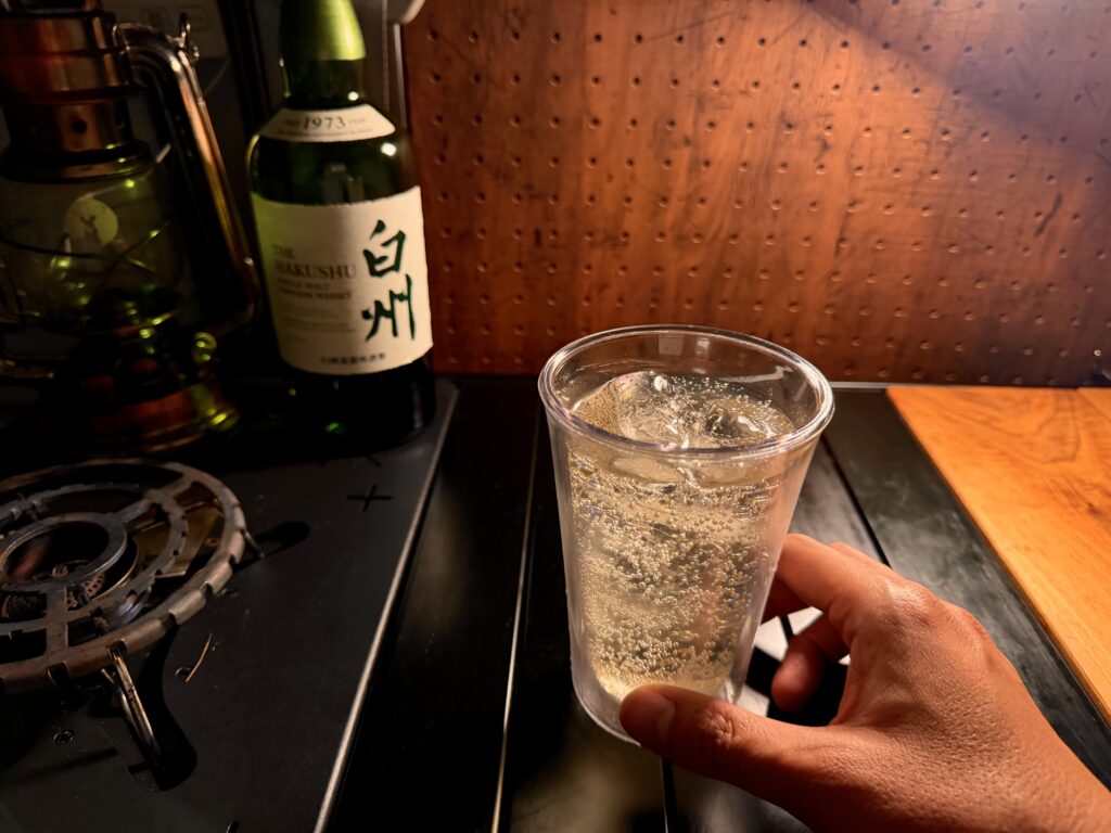 車中泊仕様のエブリイ車内で、白州のボトルを横に置き、ハイボールを手に持っている様子