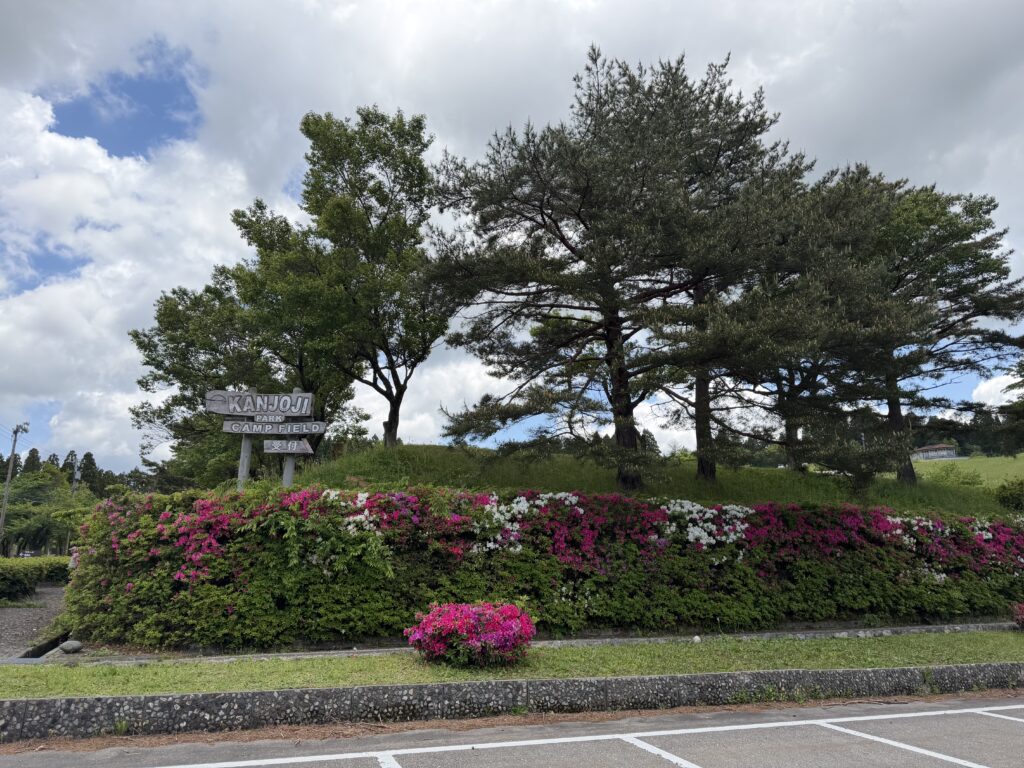 ツツジが咲く閑乗寺公園キャンプ場の入口看板と、周囲の木々や空が写っている到着時の風景