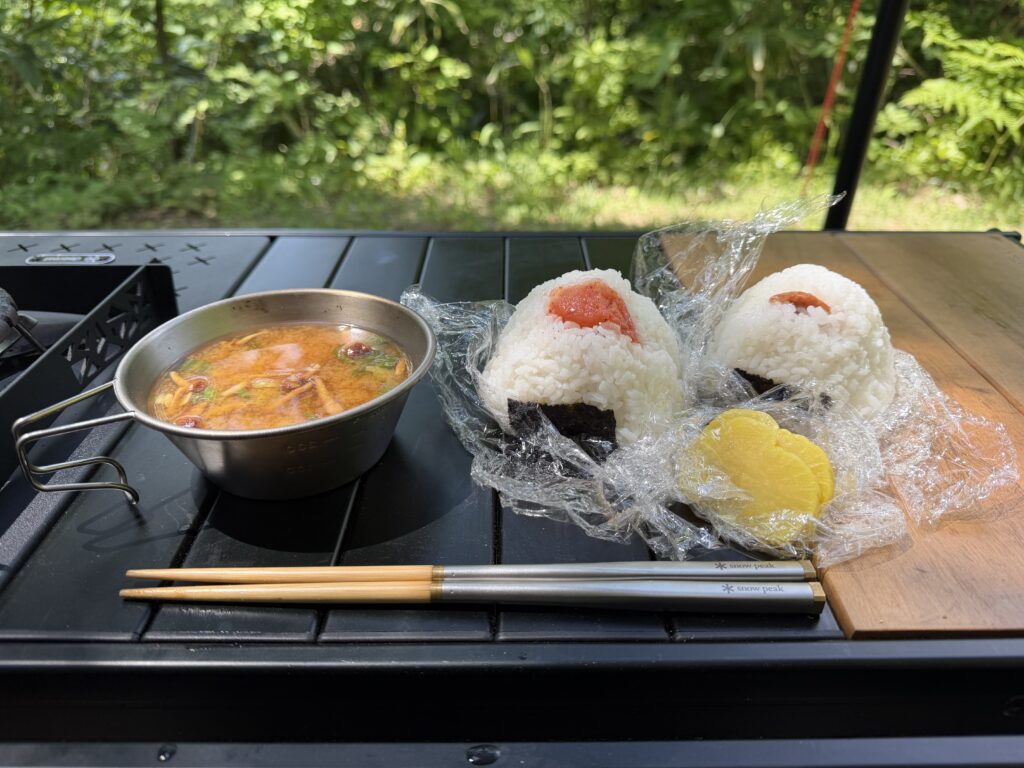 キャンプ用テーブルの上に、家でにぎってきた梅干しと明太子のおにぎり、インスタント味噌汁、たくあんを並べた昼ごはんの様子