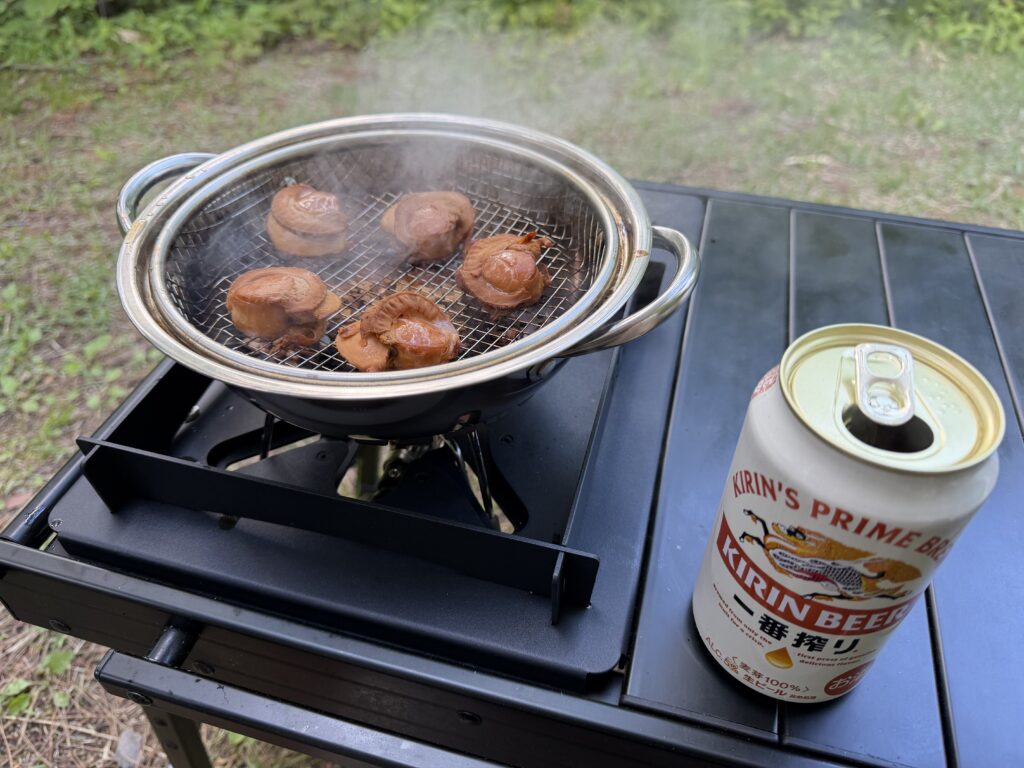 鍋の網の上で茶色く色づいた帆立の燻製と、横に置いた缶ビールが写っている様子。鍋からはまだ煙が上がっている。