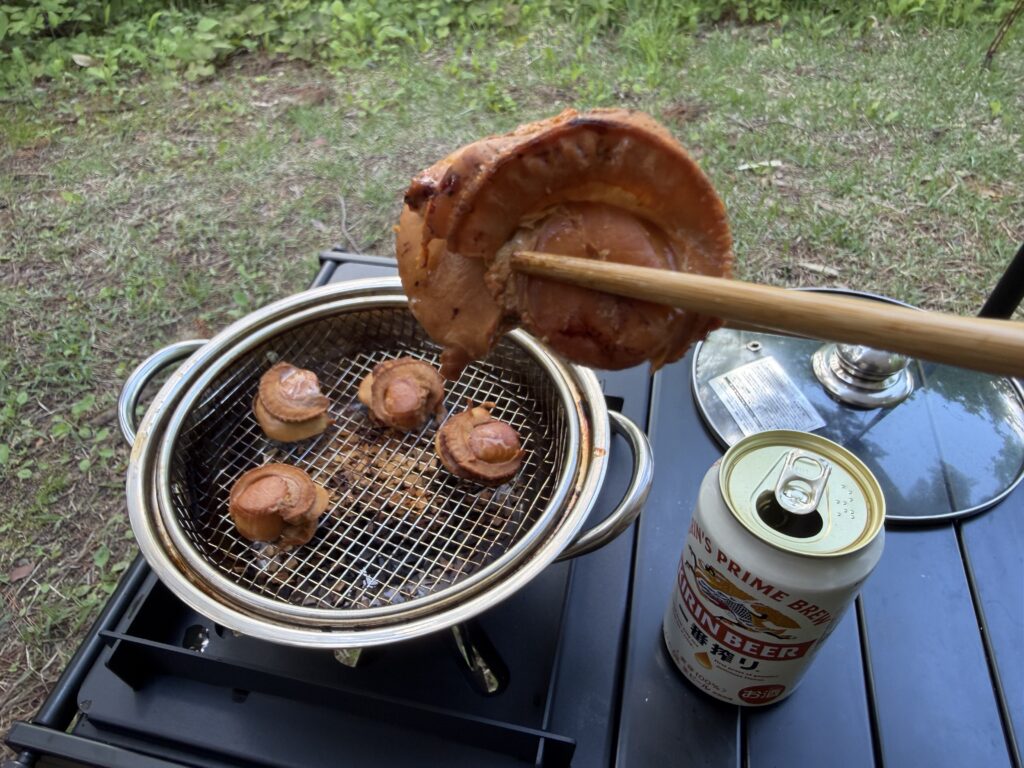 茶色く燻された帆立の燻製を箸で持ち上げ、燻製鍋と缶ビールを背景に口へ運ぶ直前の様子
