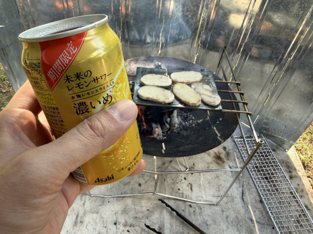 焚き火の上の鉄板で長いもを焼きながら、手前に期間限定の濃いめ未来のレモンサワーの缶を持っている様子