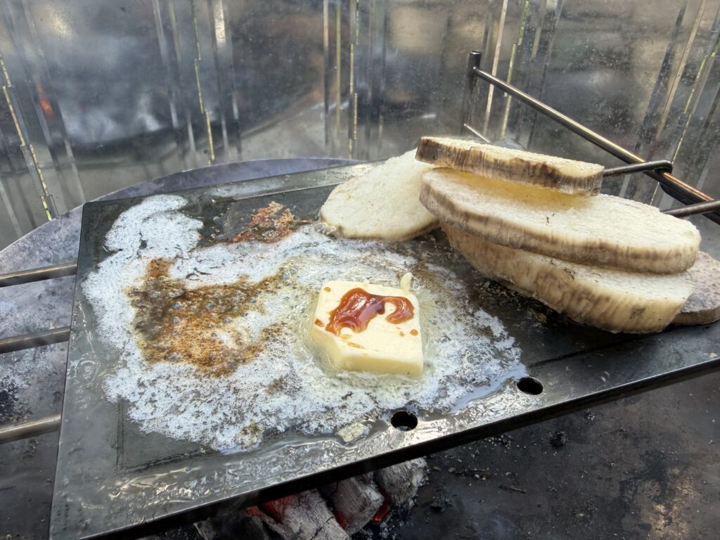 焚き火の上の鉄板で焼いた長いもを端に寄せ、溶けたバターと醤油の香りが立ち込めている様子