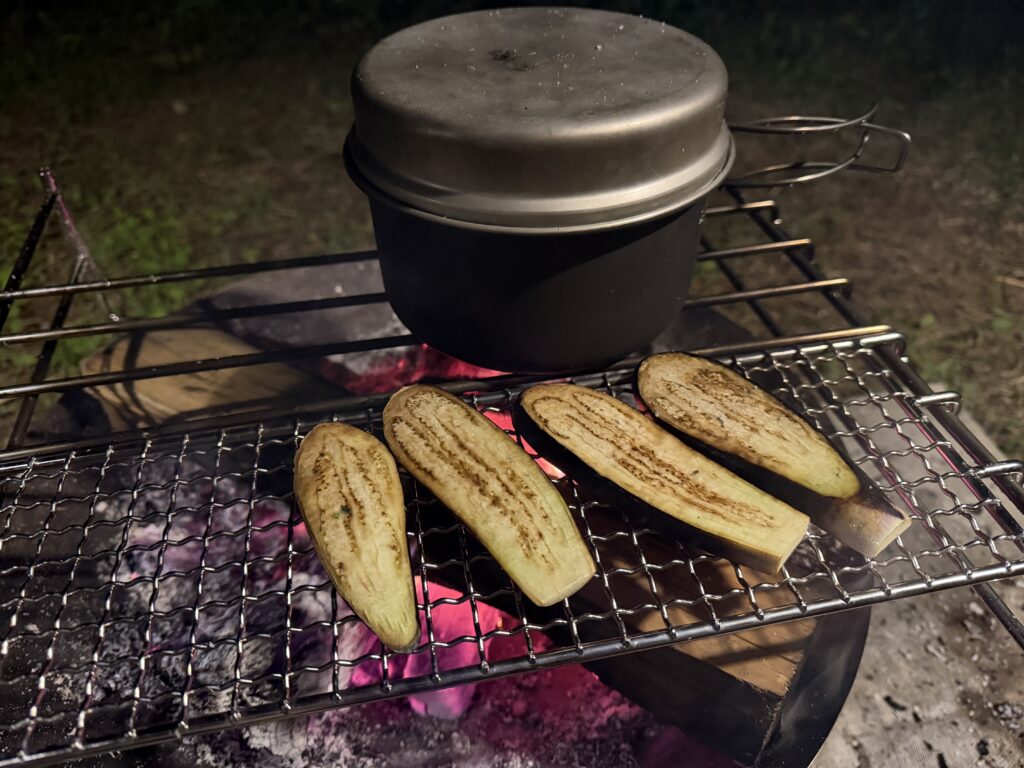 焚き火の上でクッカーを使って水蒸気炊飯をしながら、網の上でスライスしたナスを焼いている様子