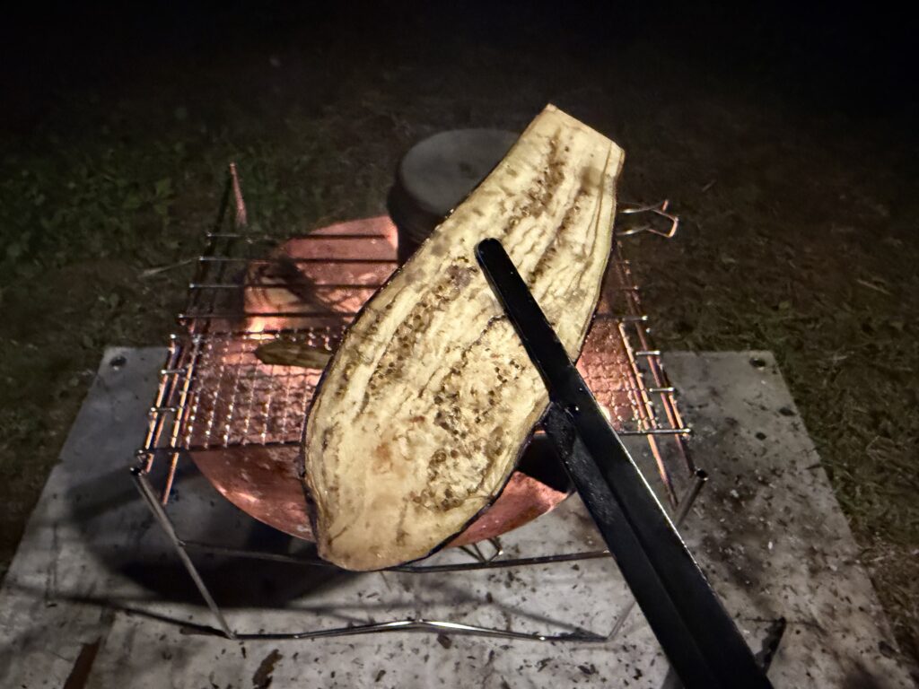 焚き火で焼いたスライスナスをトングで持ち上げ、焼き具合を確認している様子