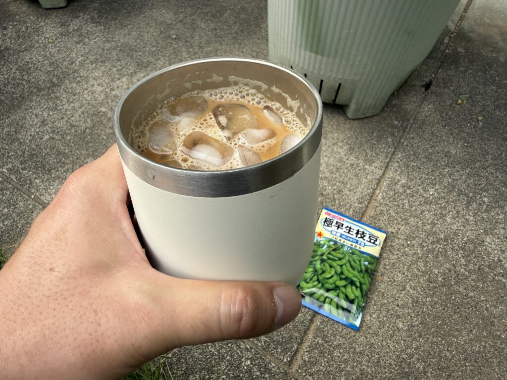 アイスコーヒーを持ちながら、奥に枝豆の種袋が置かれている様子