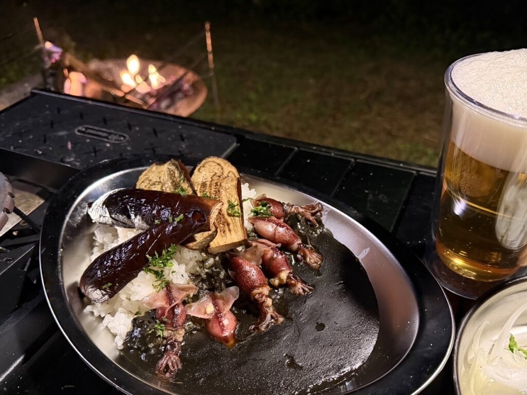 焚き火を背景に、ホタルイカと焼きナスをのせたカレーとビールを並べたキャンプ飯