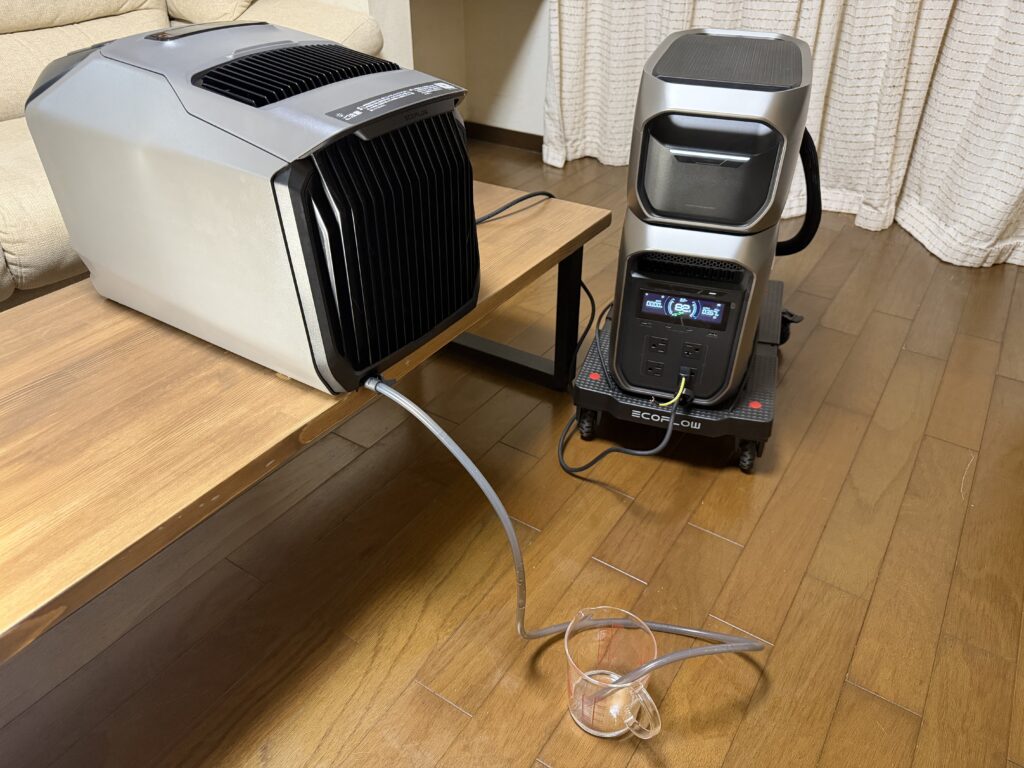EcoFlow WAVE 3の排水口にホースを接続し、ホースの先を計量カップに入れて排水している様子。隣にポータブル電源が置かれている。