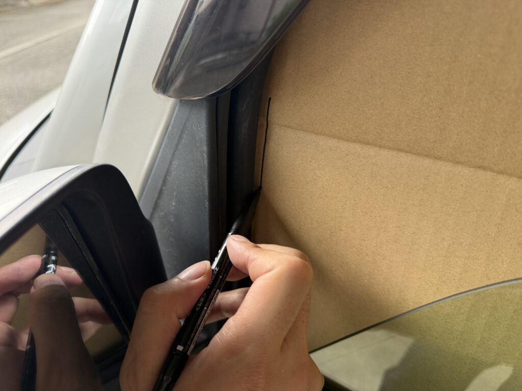 助手席窓に当てたダンボール型紙の余分な部分に、ペンでカットラインを引いている様子。