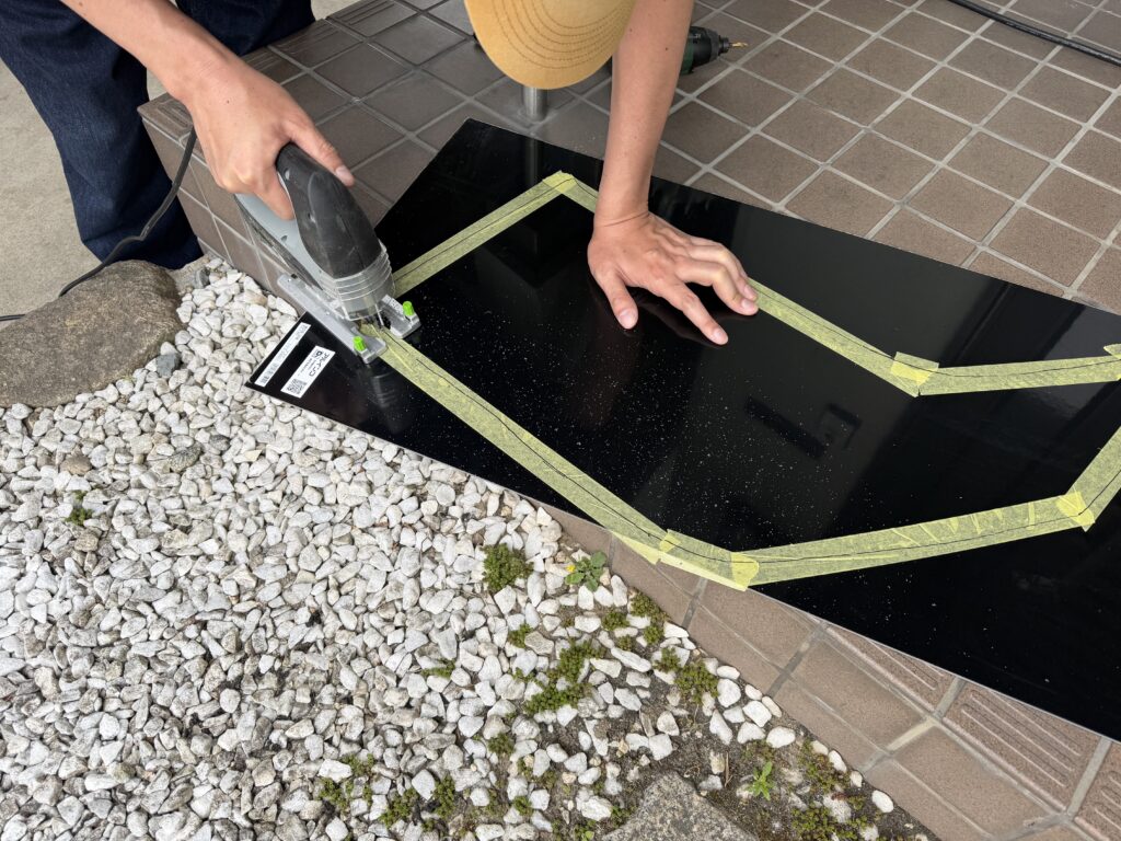 黒いアルミ複合板に写した窓パネルのラインに沿って、ジグソーで外形をカットしている様子