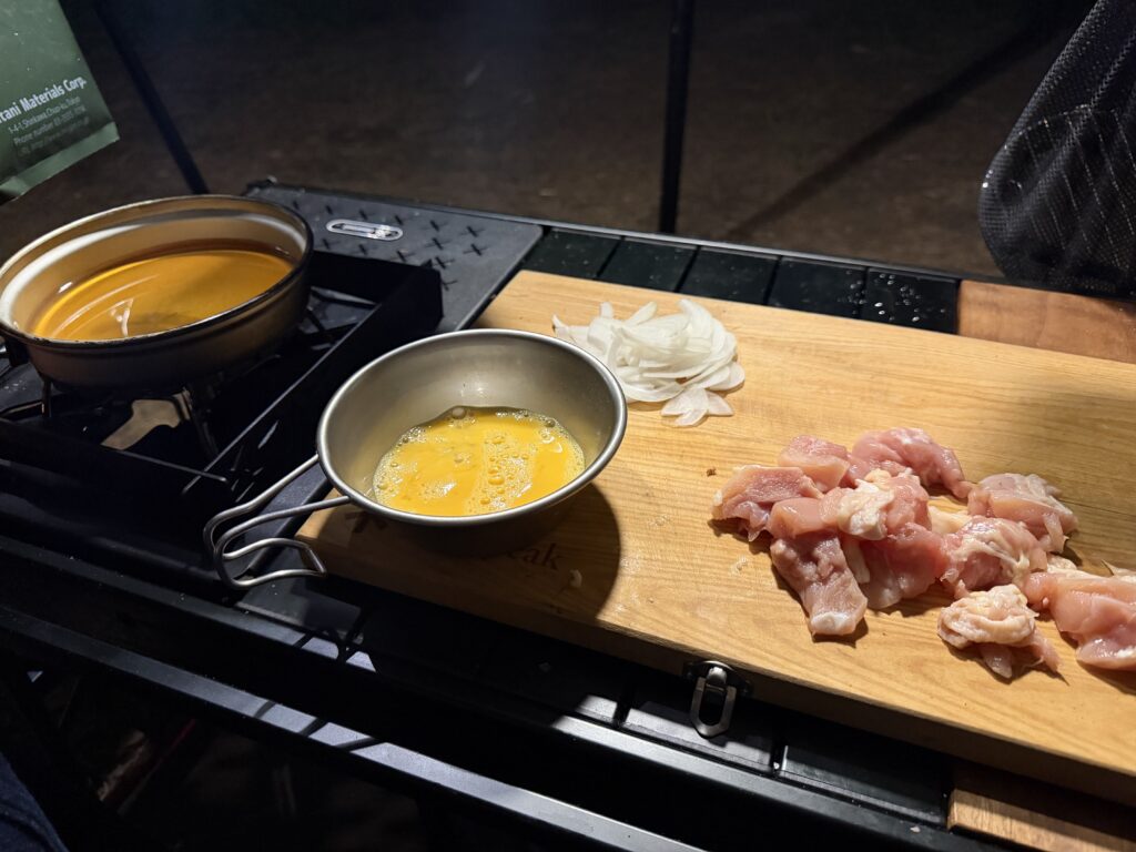 一口大に切った鶏もも肉、薄切りにした玉ねぎ、シェラカップに入れた溶き卵とクッカーにめんつゆと水を入れた様子