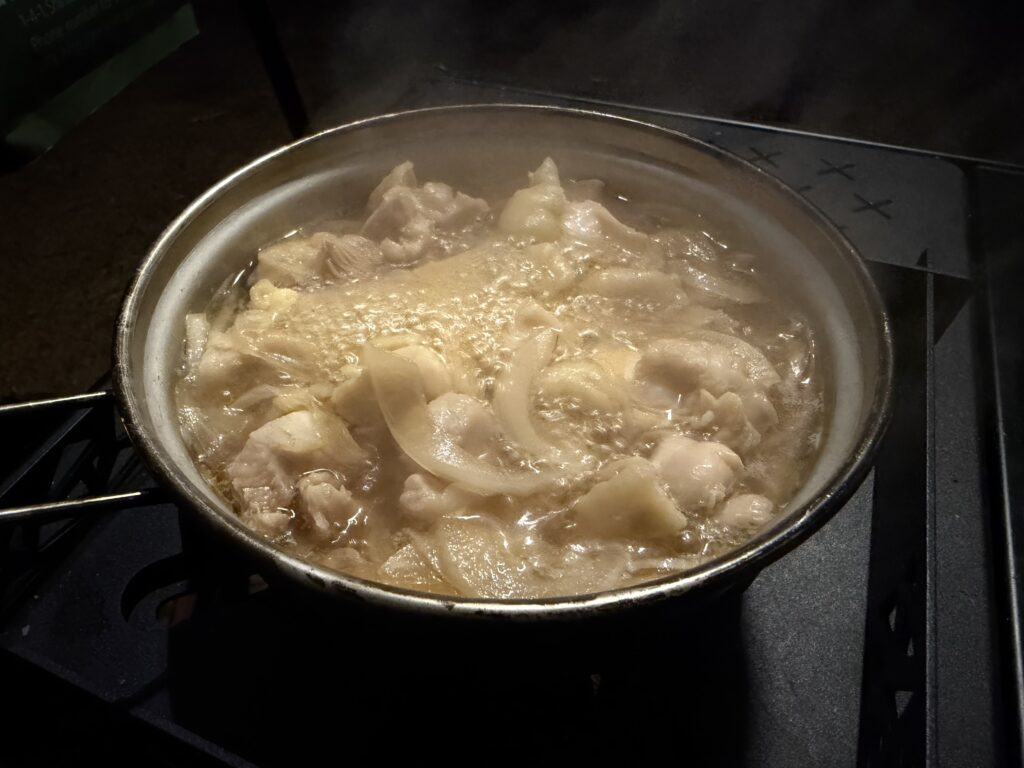 キャンプ用クッカーで鶏もも肉と玉ねぎを水とめんつゆで煮ている様子