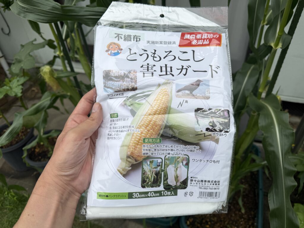 とうもろこしの株の前で手に持った不織布製とうもろこし害虫ガードのパッケージ