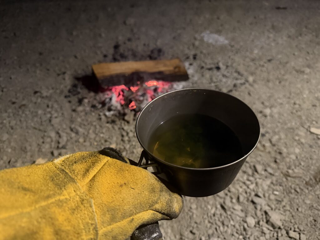 直火の残った火を眺めながら、シェラカップに入れたインスタントのしじみ汁を飲んでいる様子
