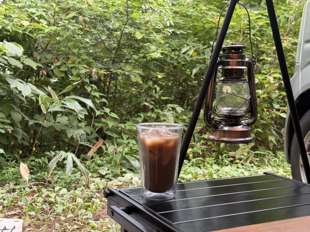 木々に囲まれたサイトのテーブルに置いたアイスコーヒーとランタン