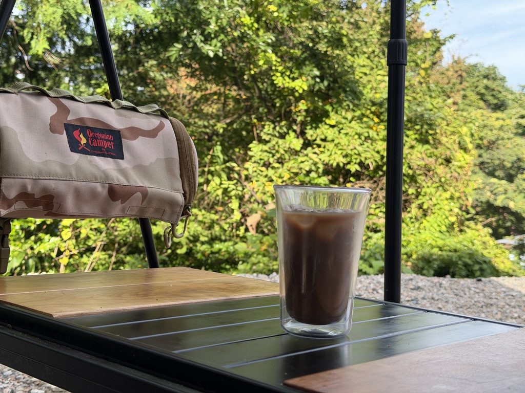 朝の木々を背景に、タープ下のテーブルに置いたアイスコーヒー