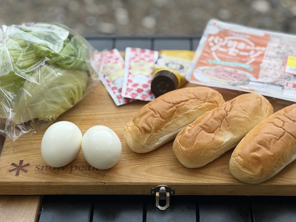 朝食用のパン3個とゆで卵、ソーセージ、レタス、調味料を並べた様子